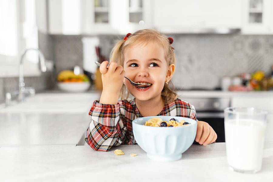 Beautiful little girl having breakfast with cereal, milk and blueberry in kitchen Photograph by Bulatovic