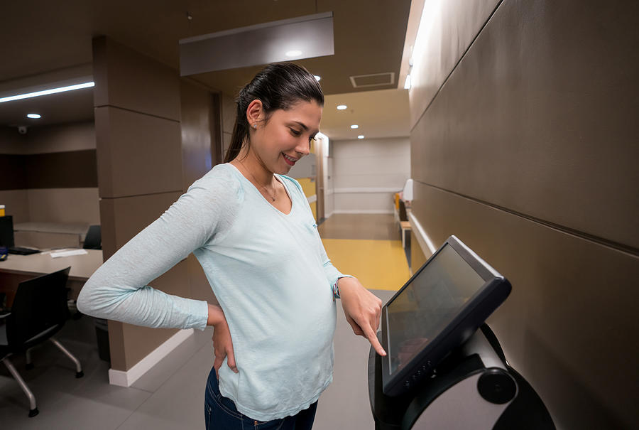 Beautiful pregnant woman requesting a number on screen at the hospital looking very happy Photograph by Andresr