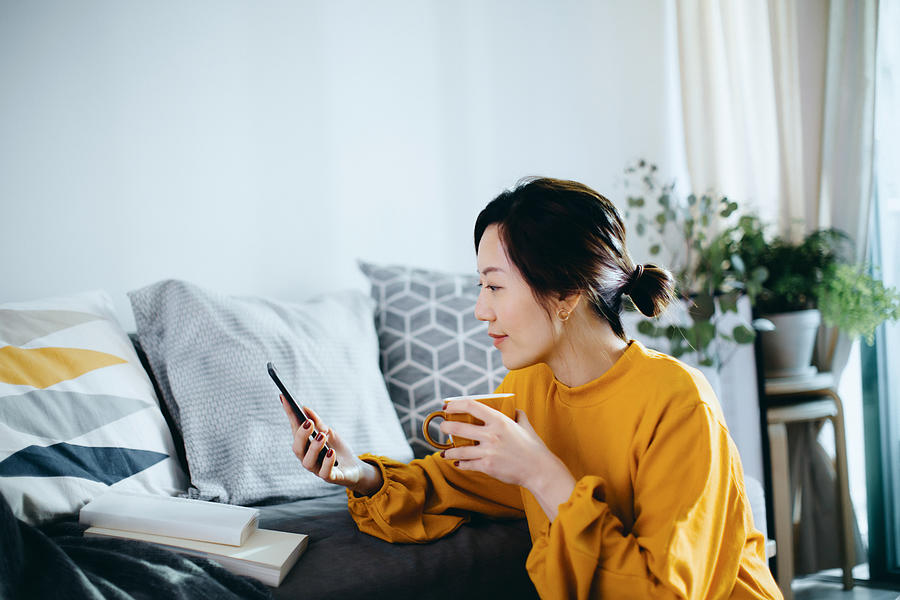 Beautiful smiling young Asian woman chilling at cozy home, sitting on the floor by the sofa, enjoying a cup of coffee and using smartphone Photograph by D3sign