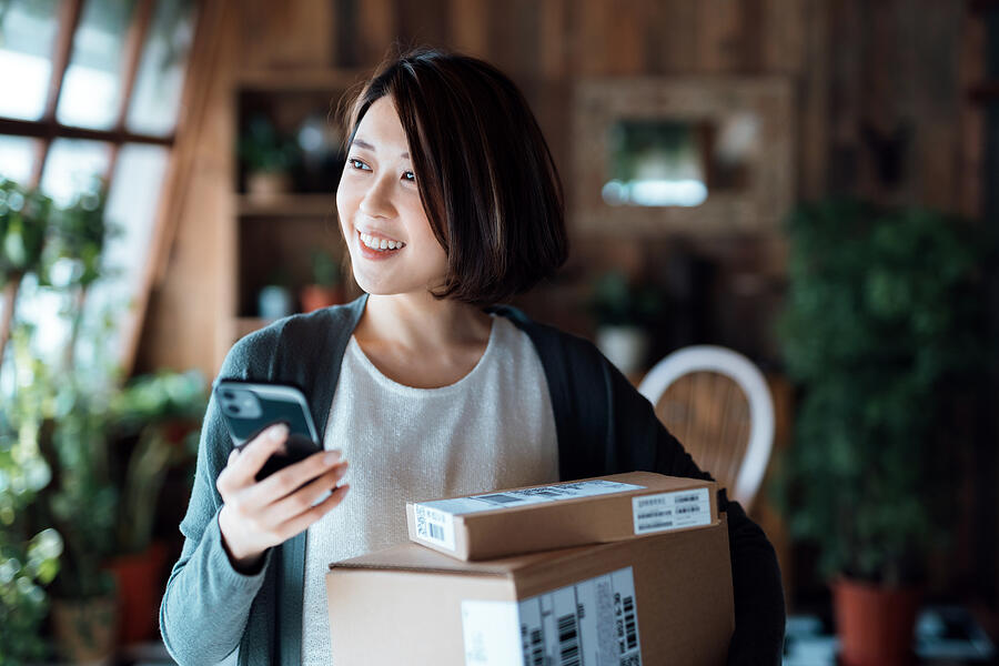Beautiful smiling young Asian woman with smartphone, receiving parcels with home delivery service at home. Online shopping, mobile payment. Enjoyable shopping experience Photograph by AsiaVision