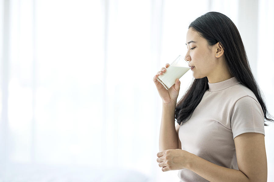 Beautiful Teenage Asian woman in casual clothing holding a glass of milk ready to drink by the window in the bedroom at home after wake up in the morning. Healthy lifestyle, Relaxing at home concept. Photograph by Nitat Termmee