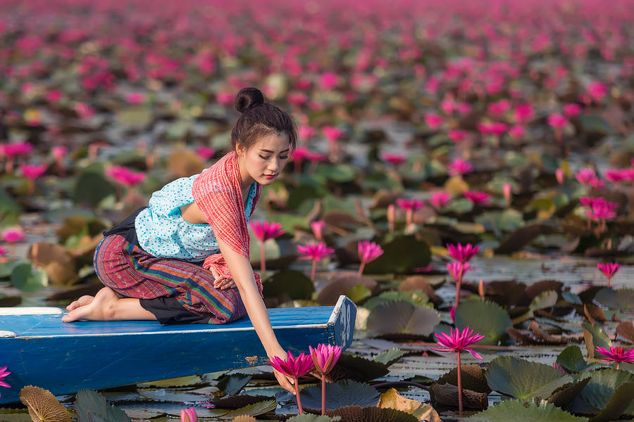 Beautiful Thai lady in Thai dress sitting on boat is holding lotus in the river. Photograph by Visoot Uthairam