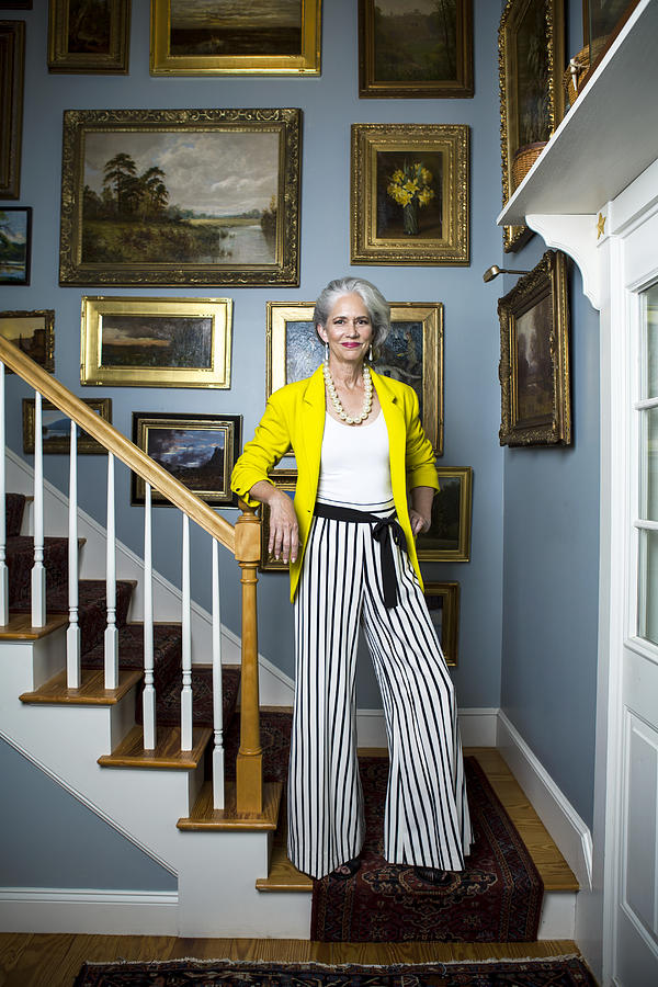 Beautiful woman in a fashionable outfit with silvery, grey hair standing in a staircase with golden framed oil painting on the wall. Photograph by Andreas Kuehn