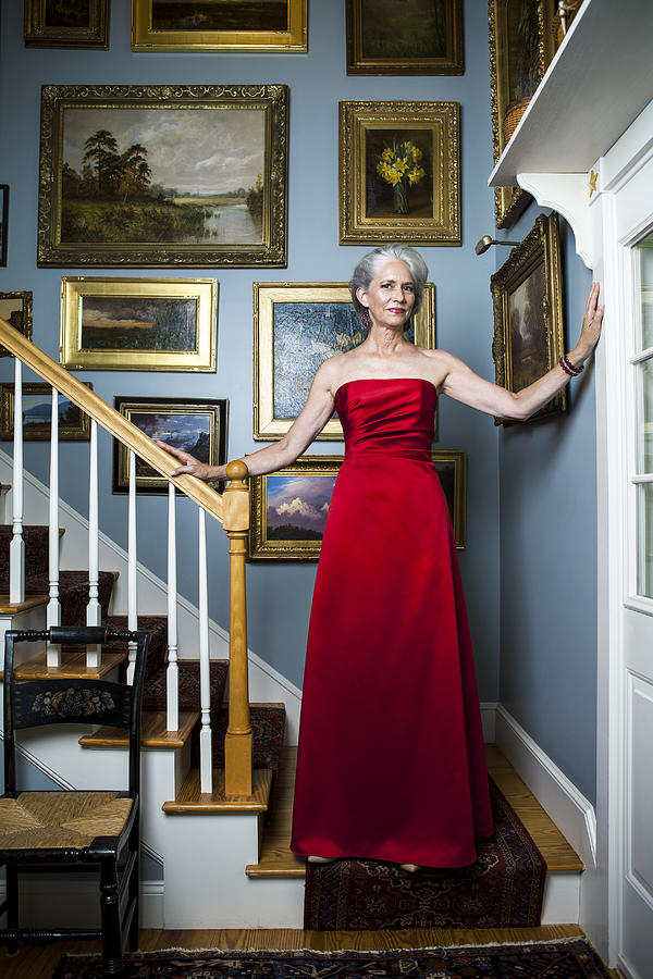 Beautiful woman in her late fifties with silvery, grey hair wearing a glamorous, red evening gown while standing in a staircase in front of golden framed oil paintings. Photograph by Andreas Kuehn