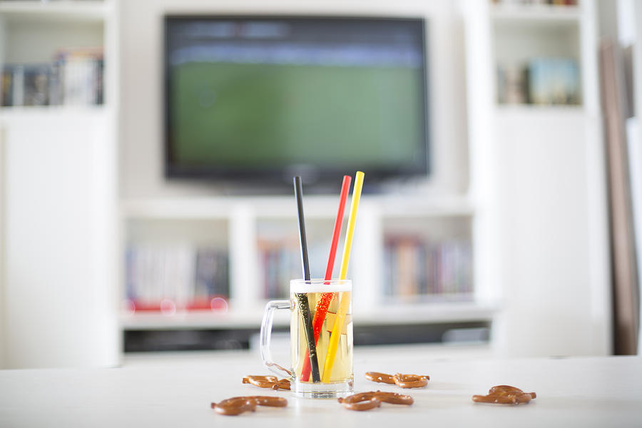 Beer mug with three drinking straws in German national colours and pretzels in front of television with empty soccer field Photograph by Westend61