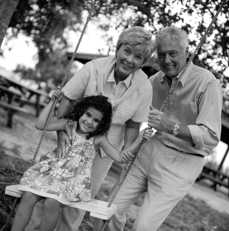 Black And White Portrait Of Grandparents At The Swings With Their Granddaughter Photograph by George Doyle