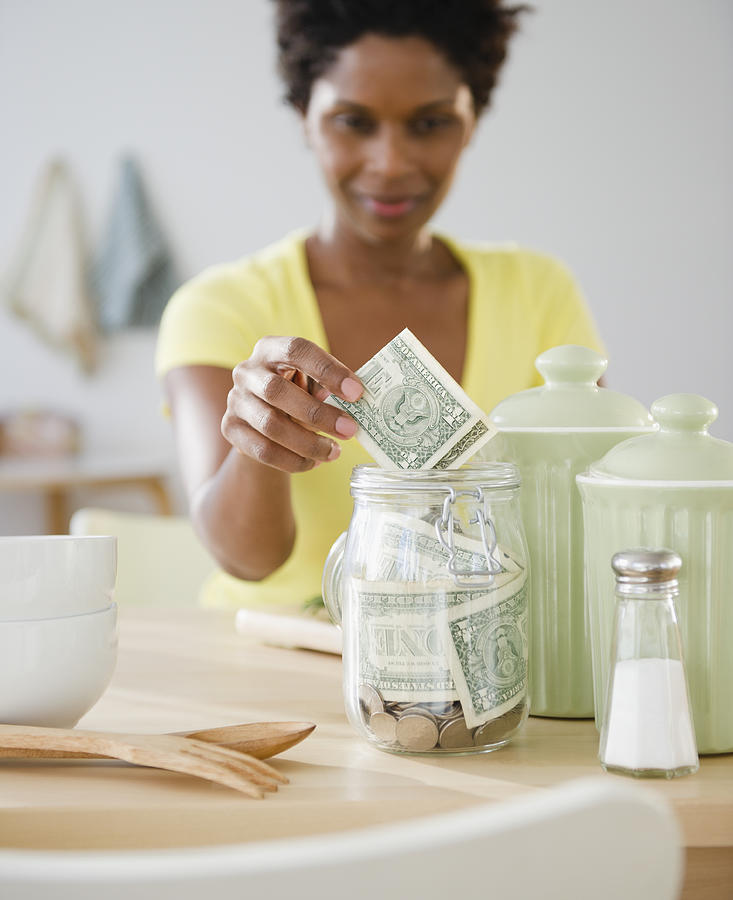 Black woman putting money in jar Photograph by JGI/Jamie Grill