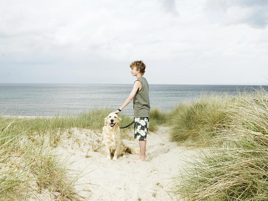 Boy (10-12) standing in sand dunes holding dog on lead Photograph by Soren Hald