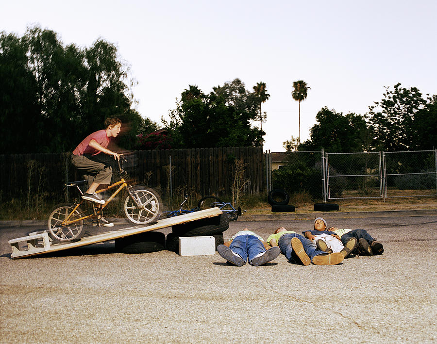 Boy (11-15) riding bicycle off jump over friends (blurred motion) Photograph by Sean Murphy