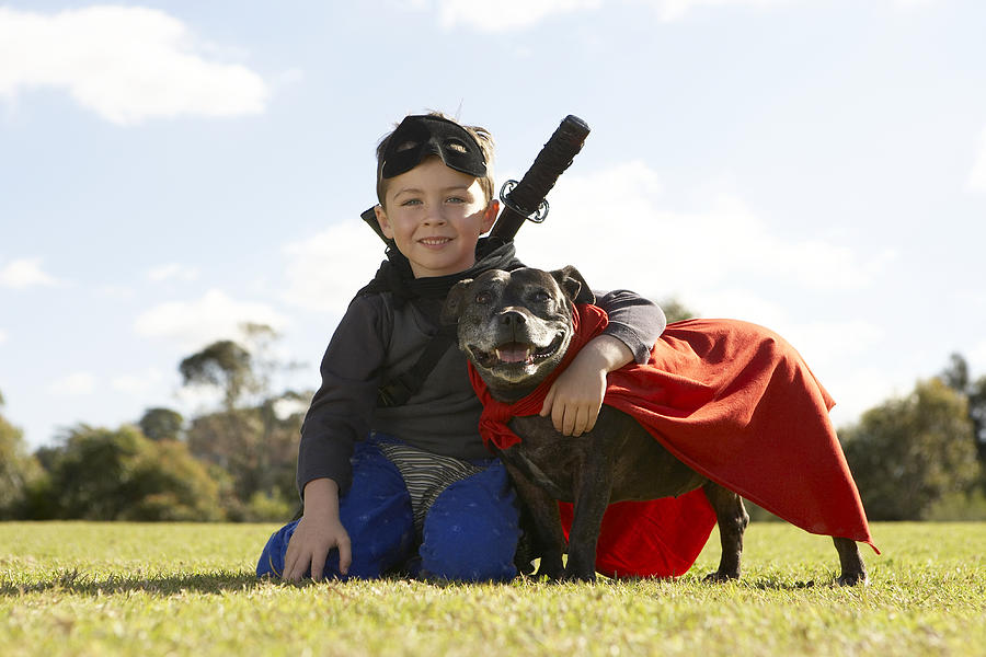 Boy (6-7) wearing costume, sitting with dog in park (surface level), portrait Photograph by Bec Parsons