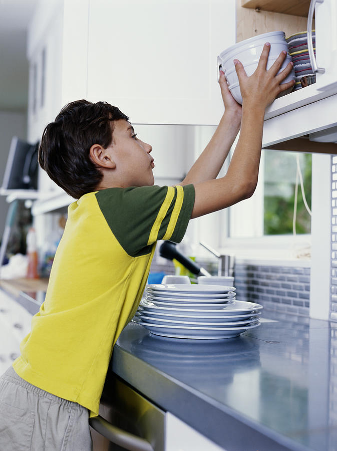 Boy (6-8) putting bowls away in kitchen cupboard, side view Photograph by Kraig Scarbinsky