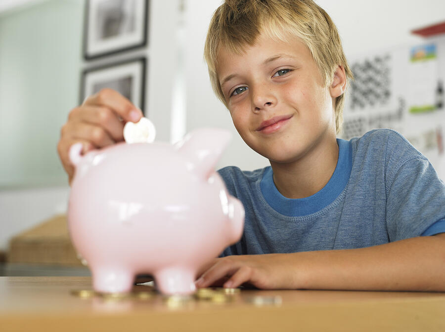 Boy (7-9) putting coin into piggy bank, smiling, portrait Photograph by Flying Colours Ltd
