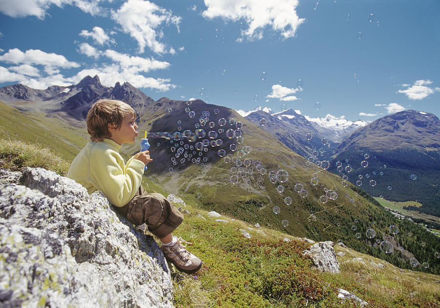 Boy (7-9) sitting on rock in mountain landscape, blowing bubble wand Photograph by Laurence Monneret