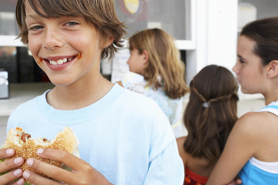 Boy (9-11) eating hamburger, portrait, three girls (9-12) queuing Photograph by Christopher Robbins