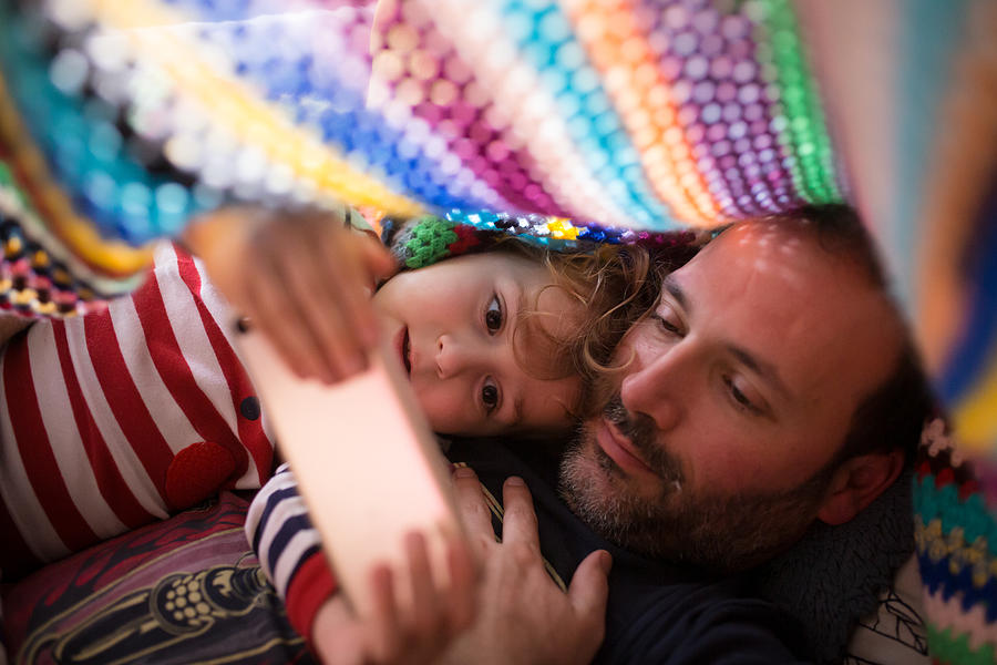 Boy and dad  watching something in a smartphone under a colorful blanket Photograph by Image taken by Mayte Torres
