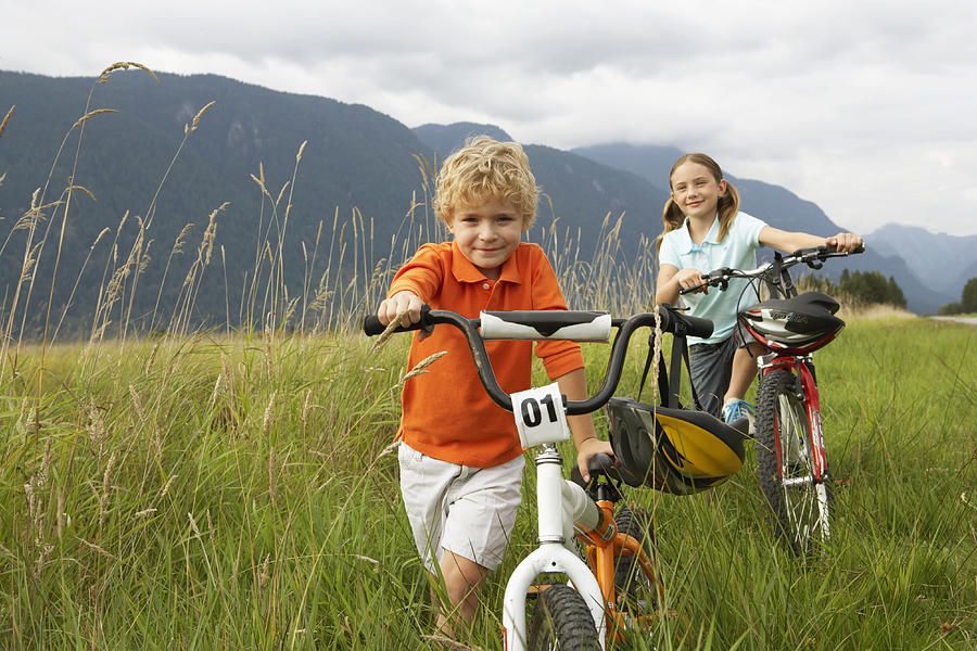 Boy and girl (5-8 years) with mountain bikes in long grass, smiling, portrait Photograph by Noel Hendrickson