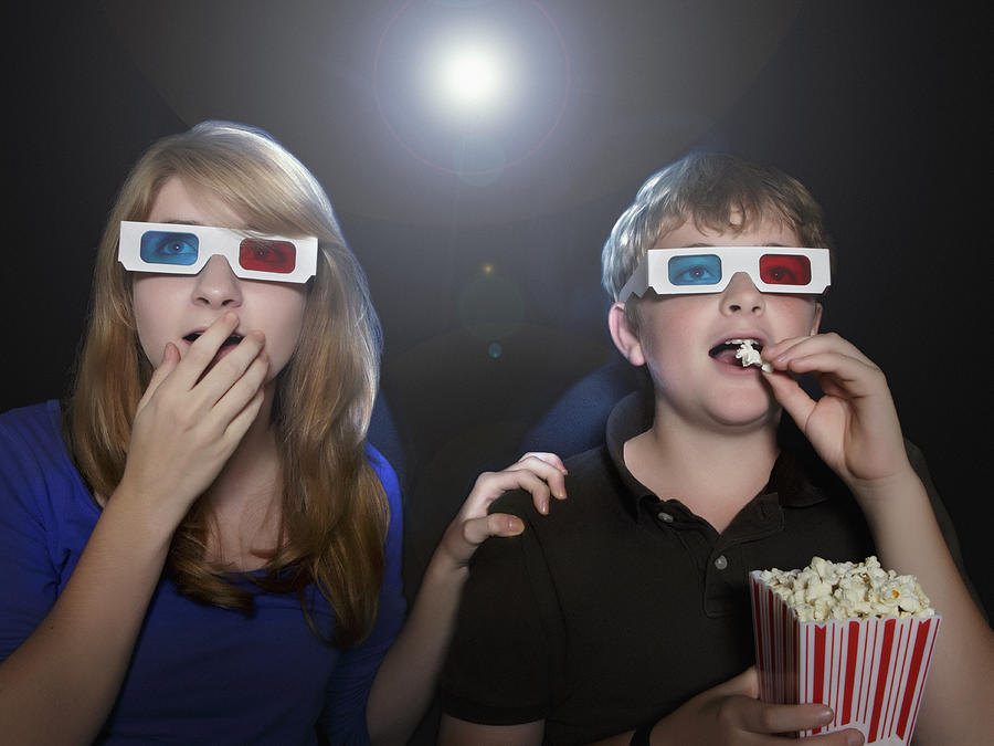 Boy and girl watching 3-D movie eating popcorn Photograph by Steven Puetzer