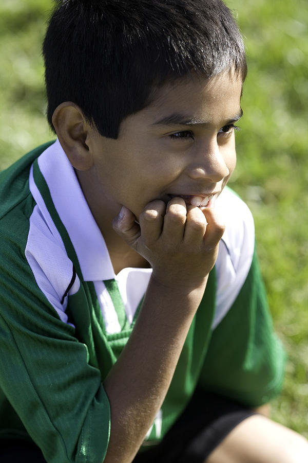 Boy in soccer uniform sitting with hand on chin, smiling, close-up Photograph by Symphonie