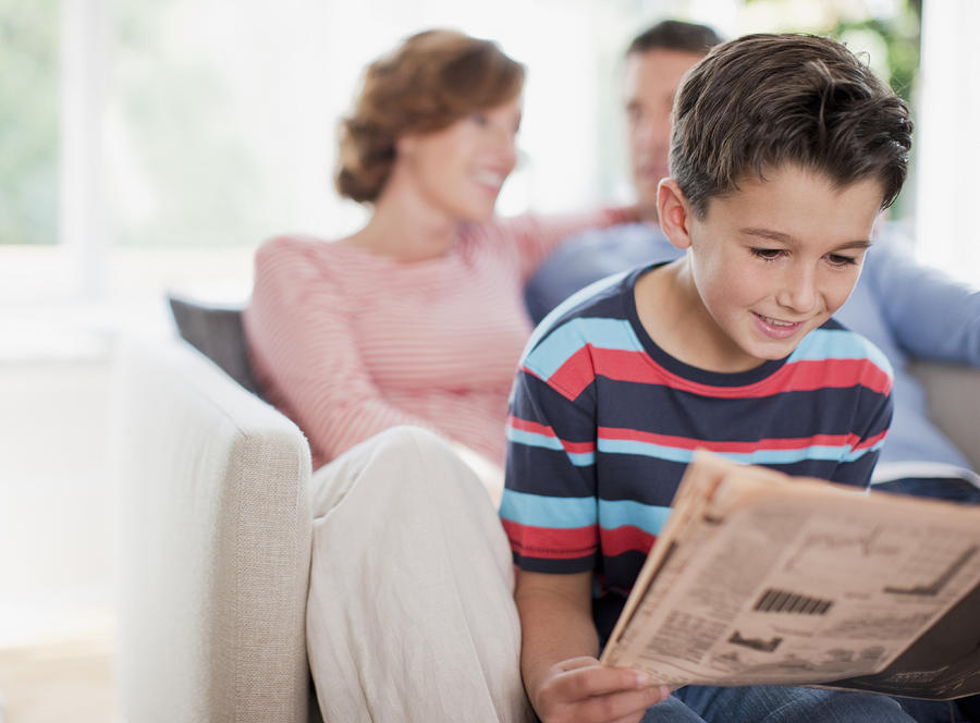 Boy reading newspaper with parents in background Photograph by Paul Bradbury