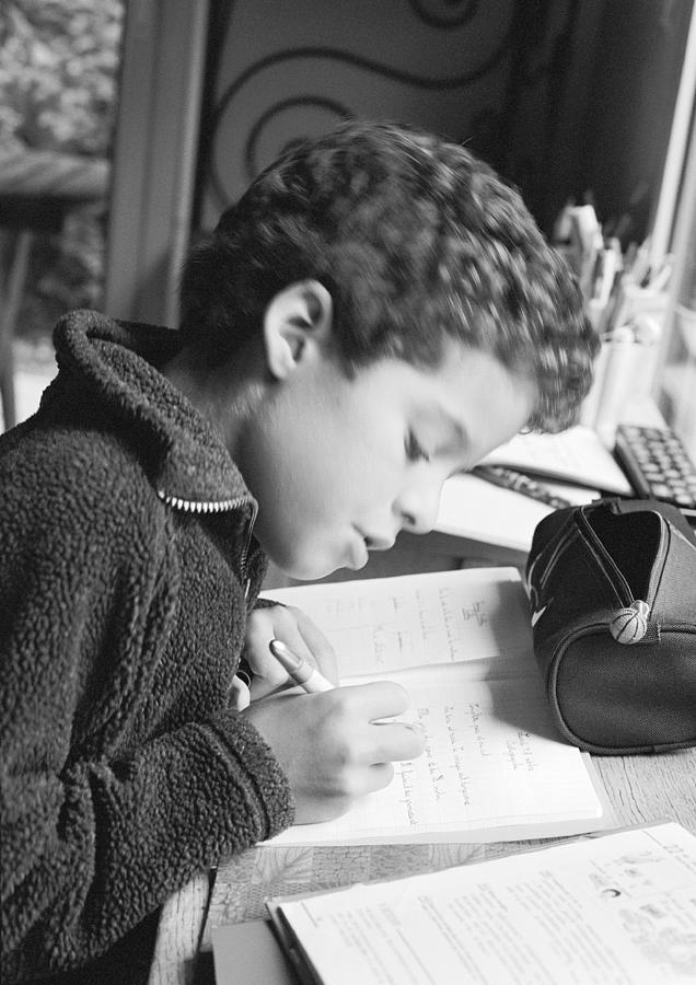 Boy sitting at desk, writing in notebook, side view, b&w Photograph by Laurence Mouton
