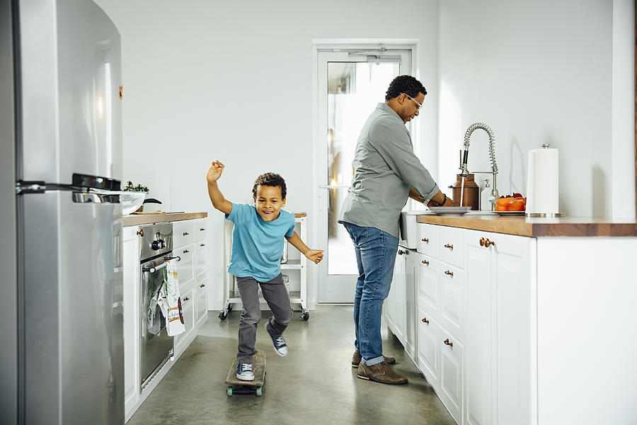 Boy skateboarding near father in kitchen Photograph by Inti St Clair