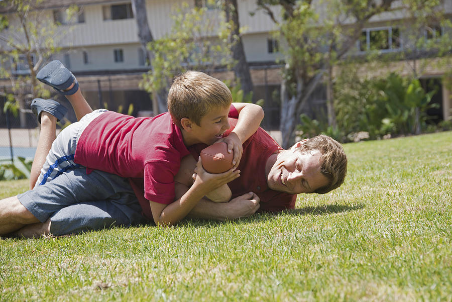 Boy tackling father playing American football in park Photograph by David Jakle