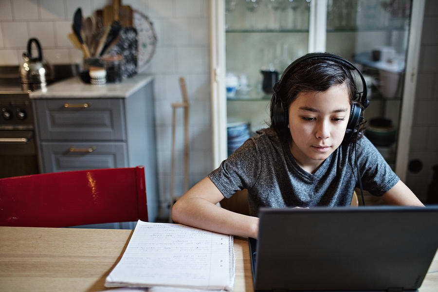Boy wearing headphones while using laptop during homework at home Photograph by Maskot