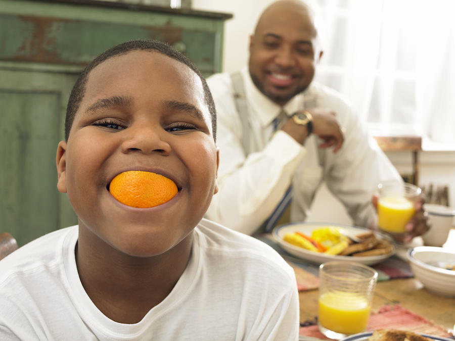 Boy With an Orange in His Mouth at Breakfast and His Father in the Background Photograph by Digital Vision.