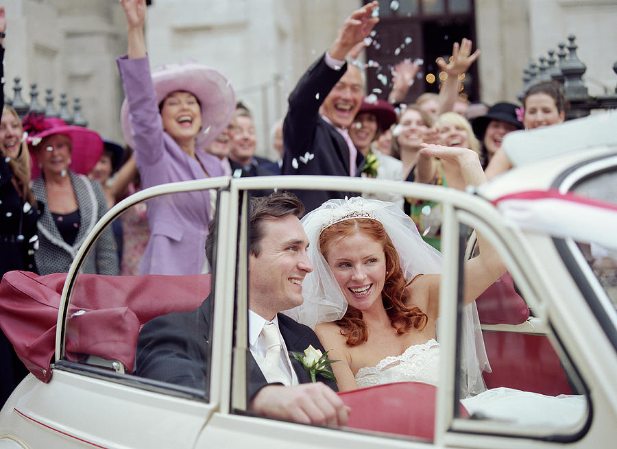 Bride and groom in convertible car, wedding party waving in background Photograph by Liz Gregg
