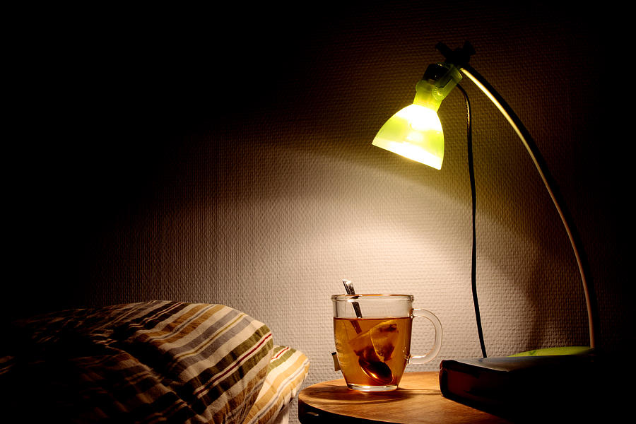 Bright bedside lamp with a clear cup of tea next to the bed Photograph by Peolsen