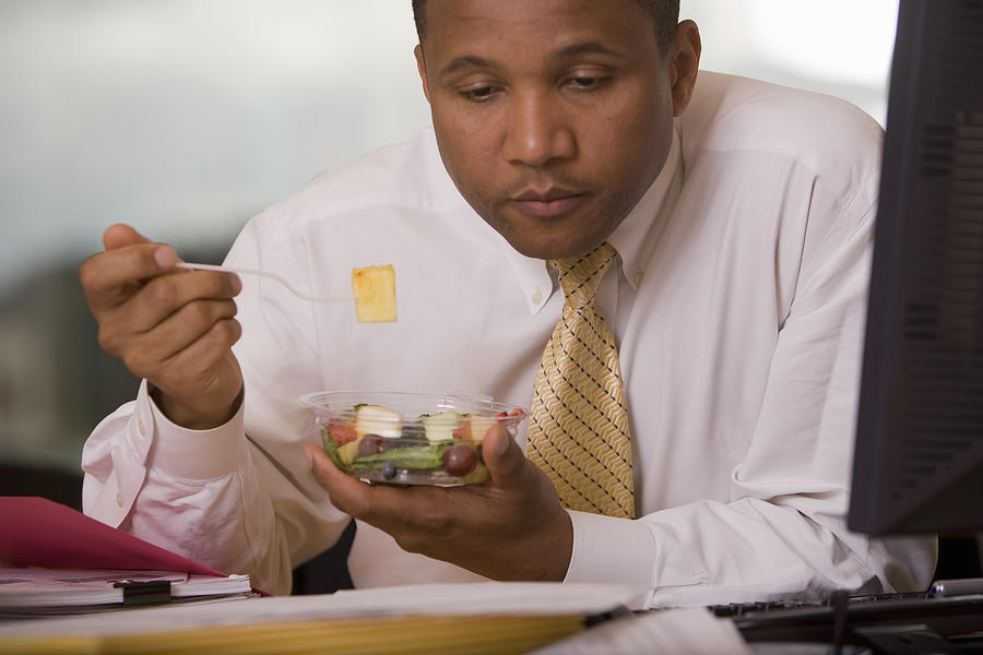 Businessman at desk eating fruit salad and reading document Photograph by John Giustina