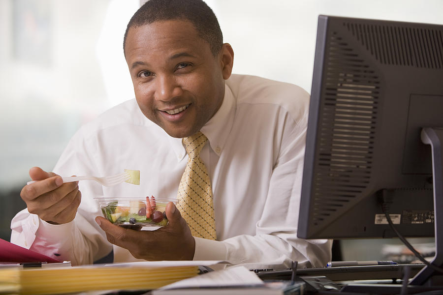 Businessman at desk eating fruit salad, smiling, portrait Photograph by John Giustina