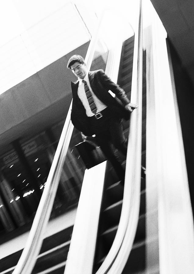 Businessman going down escalator, looking down, b&w. Photograph by Teo Lannie