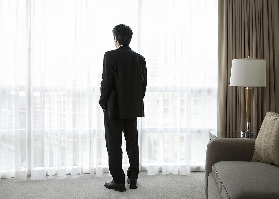 Businessman in hotel room, looking out window, rear view Photograph by Compassionate Eye Foundation/Steven Errico