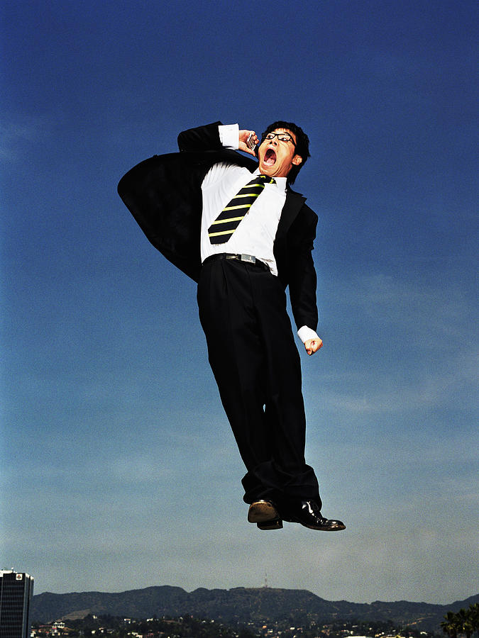Businessman jumping in air, yelling into mobile phone, low angle Photograph by Mike Powell