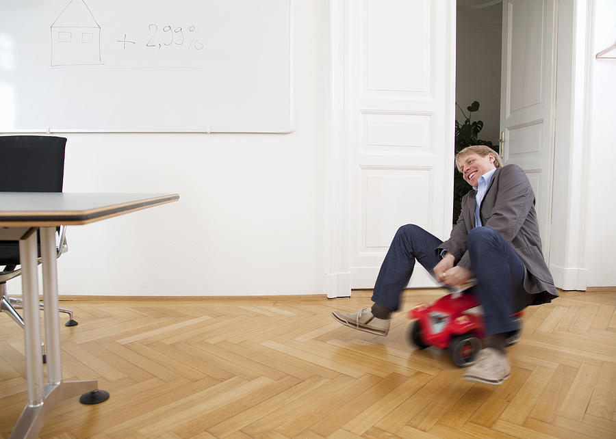 Businessman riding toy car in office Photograph by Henglein and Steets