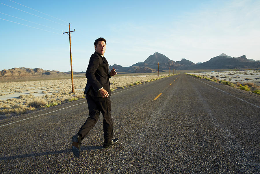 Businessman running down desert road, looking back Photograph by Andy Ryan
