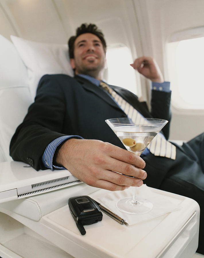 Businessman Sits in a Plane Holding a Martini, Focus on the Foreground Photograph by Digital Vision.