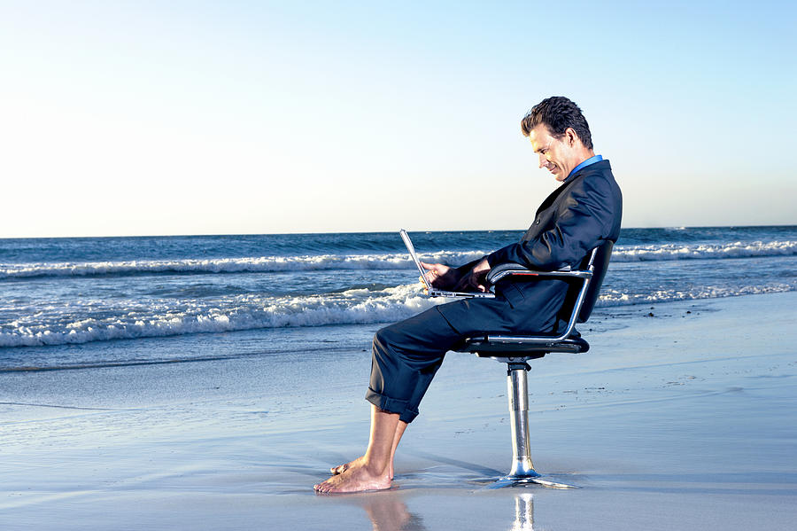 Businessman Sitting on a Beach by the Sea in a Swivel Chair and Working on His Laptop Photograph by John Cumming