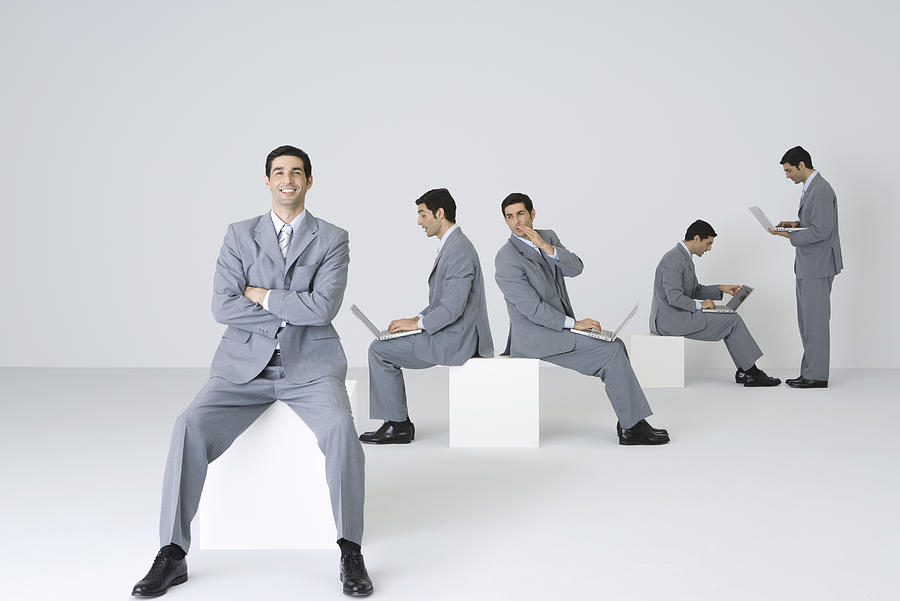 Businessman smiling at camera with arms folded while his clones use laptops in background Photograph by PhotoAlto/Ale Ventura