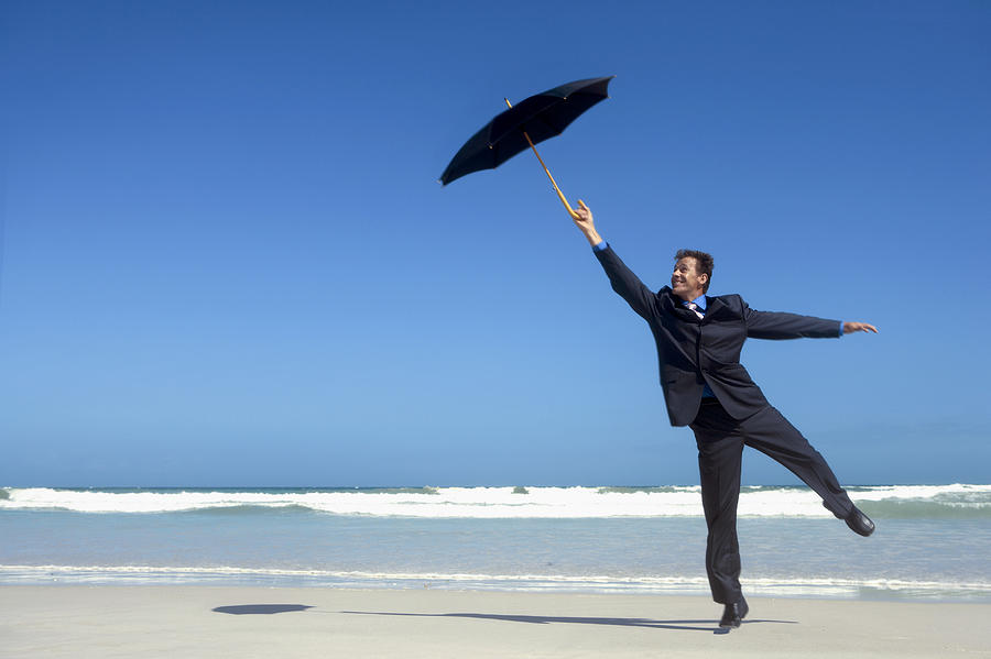 Businessman Standing on One Leg and Holding an Umbrella, on a Beach Photograph by John Cumming