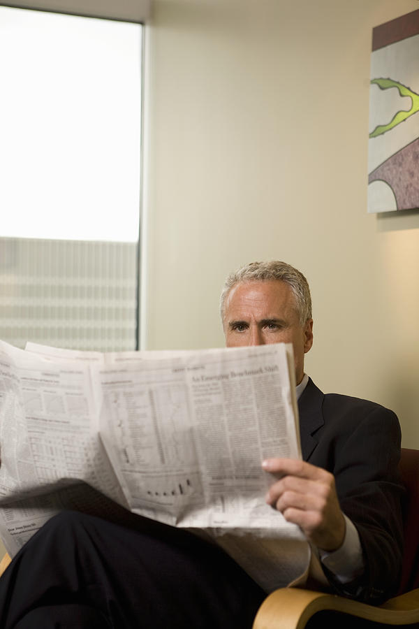 Businessman waiting in office lobby, reading newspaper Photograph by John Giustina