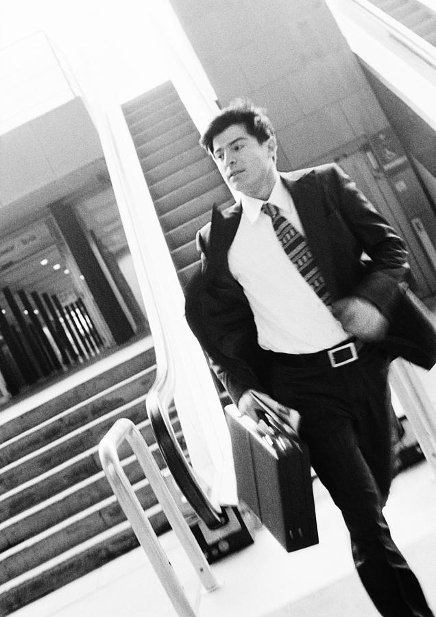 Businessman walking away from escalator, holding briefcase, blurred motion, b&w. Photograph by Teo Lannie
