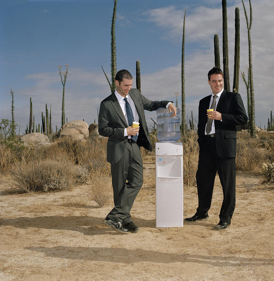 Businessmen near water cooler in desert, looking to side Photograph by Matthias Clamer