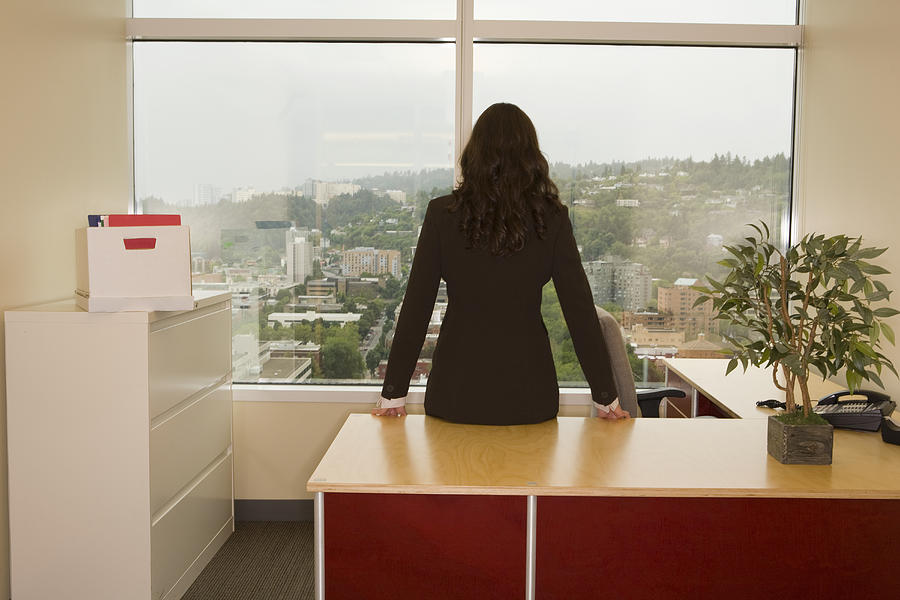 Businesswoman leaning at desk, looking through window, rear view Photograph by John Giustina