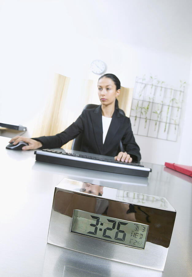 Businesswoman Sits at Desk With an Alarm Clock on it Using a Computer Mouse Photograph by B2M Productions