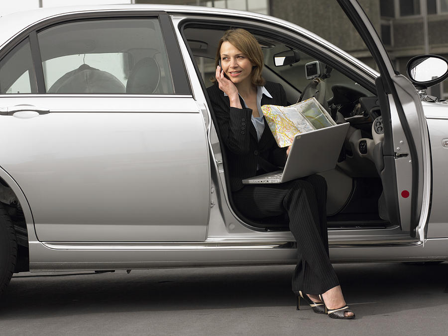 Businesswoman Sitting in Her Car Using a Mobile Phone With a Laptop and a Map Photograph by Digital Vision.