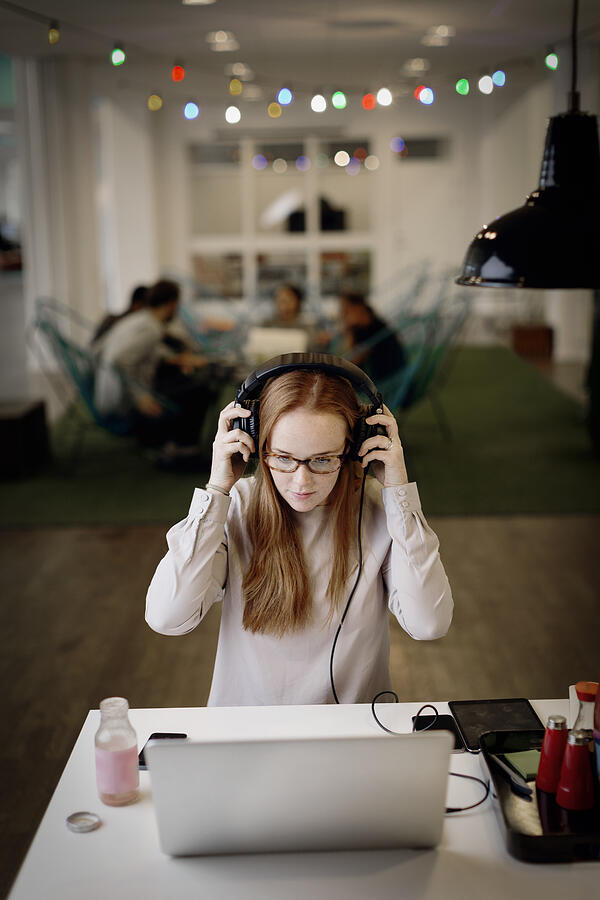 Businesswoman wearing headphones while working late on laptop in creative office Photograph by Maskot
