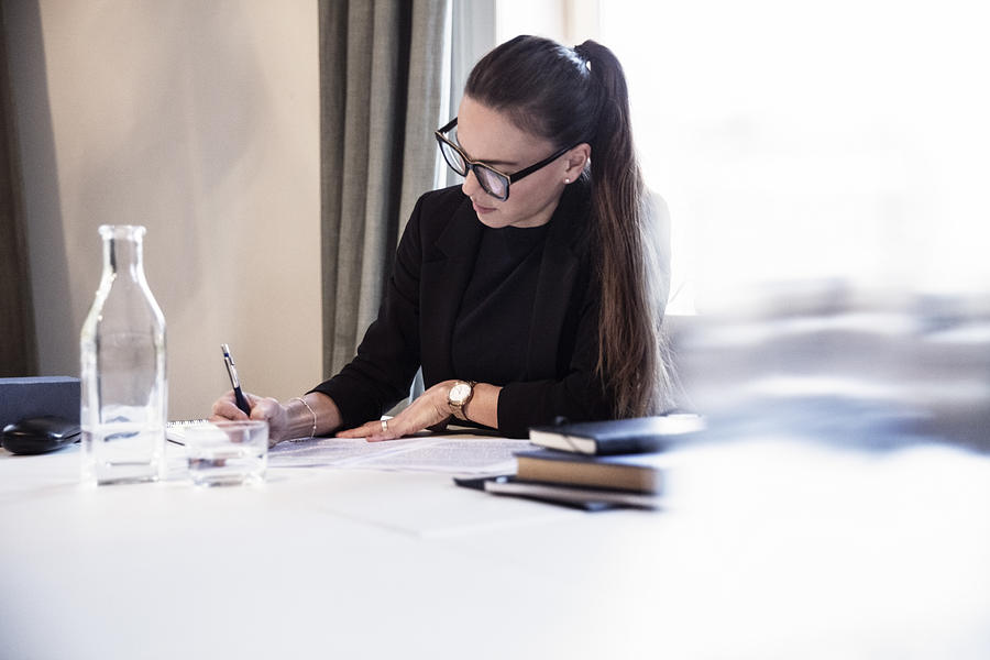 Businesswoman writing on documents while sitting by desk at law firm Photograph by Maskot Bildbyrå