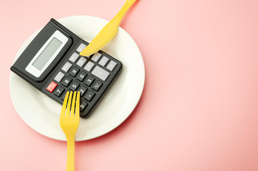 Calculate expensive food spending costs, counting calories and weight loss program concept with calculator on empty plate, yellow fork and knife isolated on pink background with copy space Photograph by Moussa81